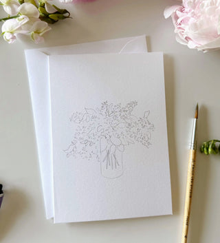 White card with floral design on a light surface with flowers and a brush.